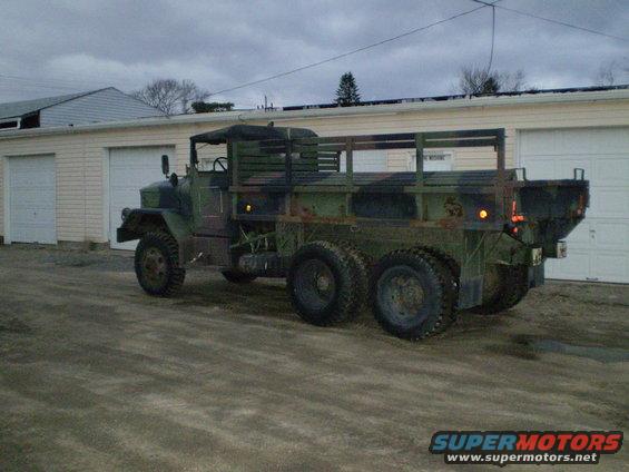 Camouflaged military truck with dual rear wheels and wooden bed, parked in front of a garage