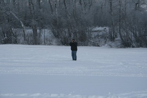 Person stands in a vast snowy field, trees in the background