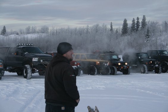 Several off-road vehicles parked in a snowy area with trees in the background. A person stands in the foreground looking away from the camera