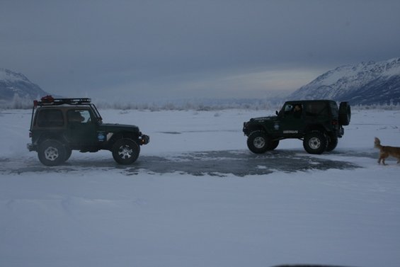 Two dark green Jeeps parked on a patch of ice in a snowy field, mountains in the distance