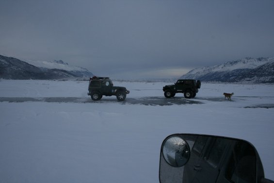 Two Jeeps driving on a frozen road with mountains in the background and a dog running nearby. Shot from inside another vehicle