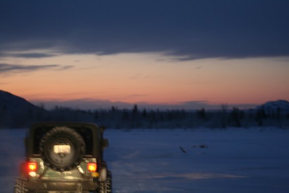 Olive green Jeep Wrangler with spare tire on rear, taillights on, in a snowy landscape at dusk