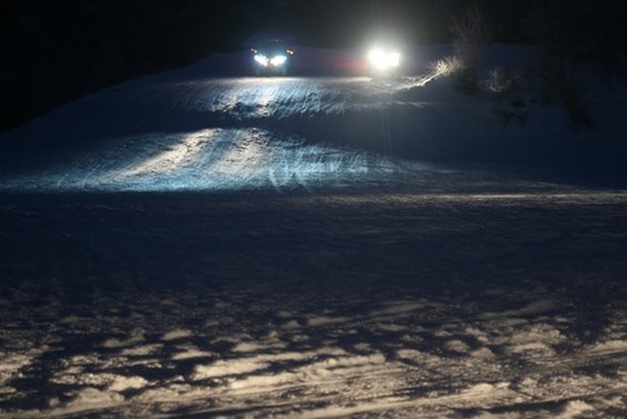Two vehicles with headlights on drive up a snow-covered hill at night
