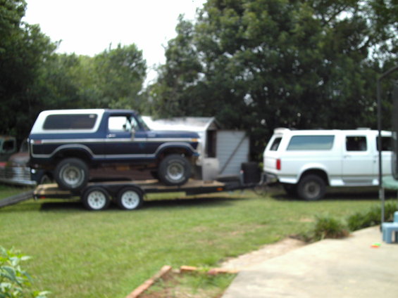 Blue and white 1978 Ford Bronco on a trailer, side view. White SUV in background