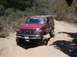 1996 Ford Bronco - A red 1996 Ford Bronco navigates a rough dirt path with one wheel dropped into a rut.