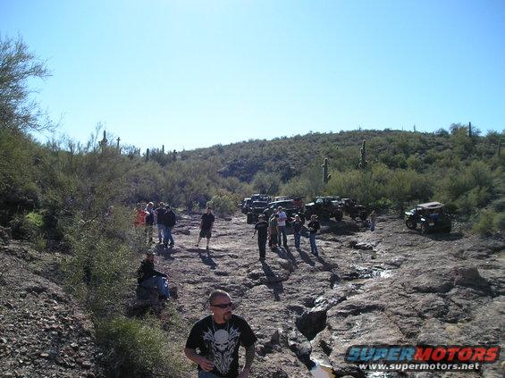 Group of people gathered around several off-road vehicles on a rocky desert trail