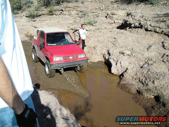Red SUV driving through a muddy trail in a desert-like area. A person stands by the open driver's door