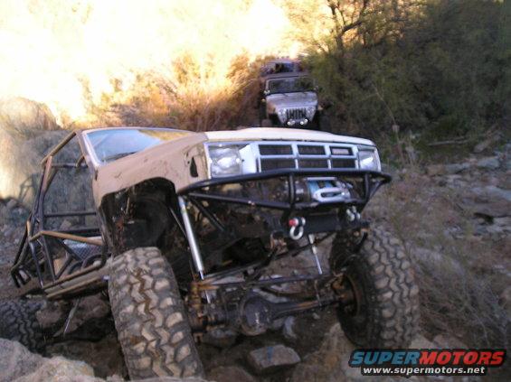 Modified pickup truck with roll cage on rocky terrain, another SUV behind it