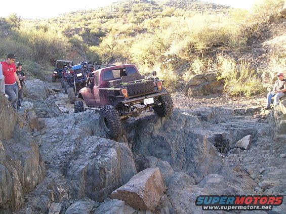 Maroon pickup truck with off-road modifications navigating a rocky trail, spare tire mounted on the back
