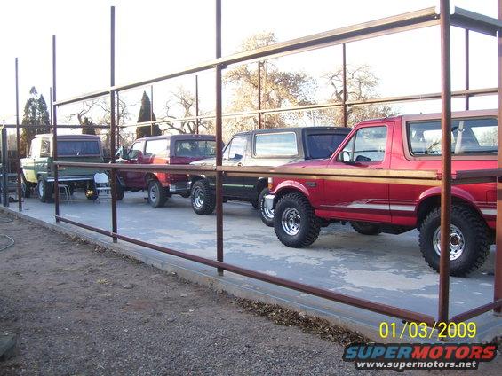 Several Ford Broncos parked in a row under a metal structure. The closest is red