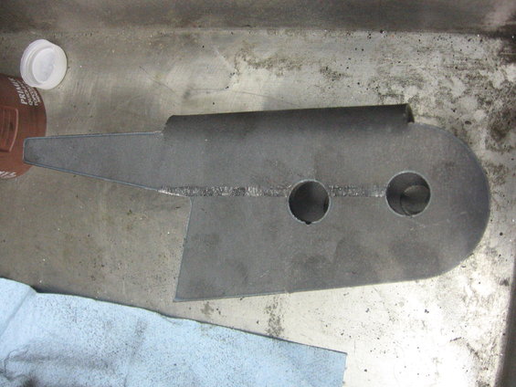 Metal bracket with two circular holes and a pointed end, resting on a surface next to a blue cloth and a bottle cap