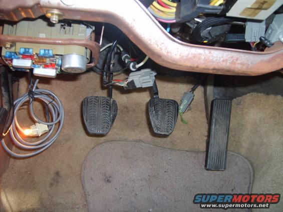 Car interior footwell showing brake and accelerator pedals, fuse box, and wiring