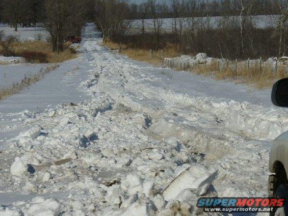 Snowy road with deep ruts and a red truck in the distance. Part of a white vehicle is visible on the right