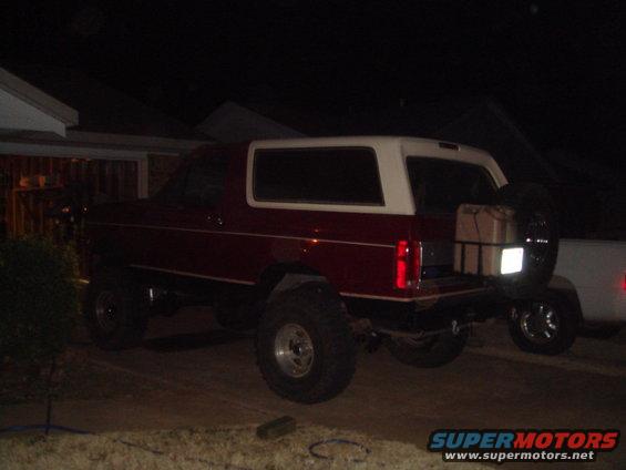 Red and white SUV parked at night, with a spare tire and gas can on the back. Another white vehicle is partially visible