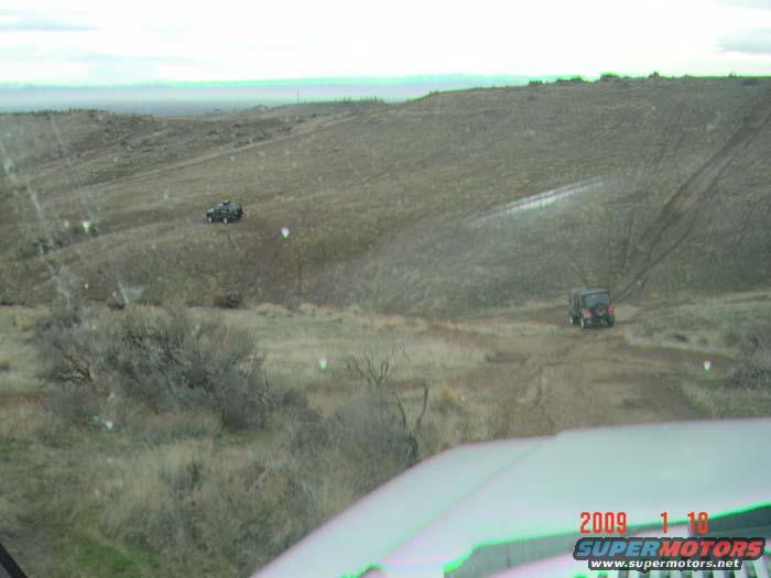 Two off-road vehicles driving on a dirt track in a hilly, barren landscape. Watermark reads 'SUPERMOTORS.NET'