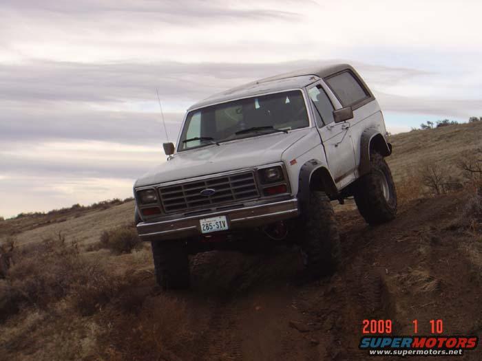 White 1980s Ford Bronco with aftermarket wheels and tires driving on a dirt hill