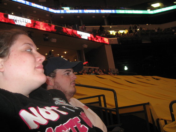 Two people watch an event in a stadium with a large yellow tarp in the foreground and a "Monster Jam" sign overhead
