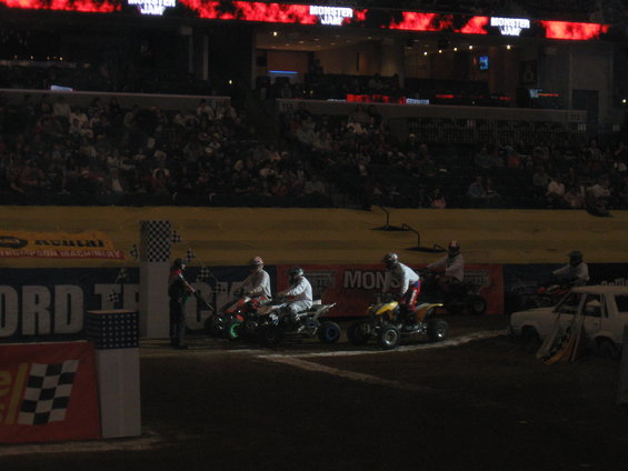 Four-wheelers lined up at a race track with checkered flags and spectators in the background