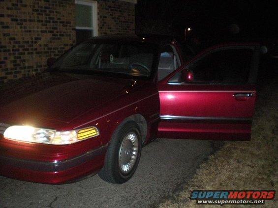 Red sedan with driver's door open, front quarter view, headlights on