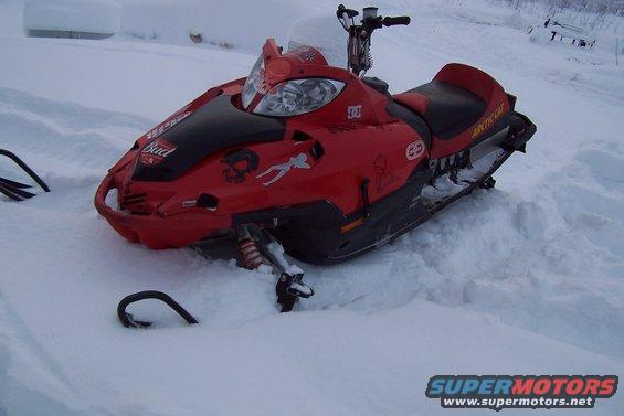 Red snowmobile with black accents and various stickers, parked in deep snow. Handlebars and part of the track are visible