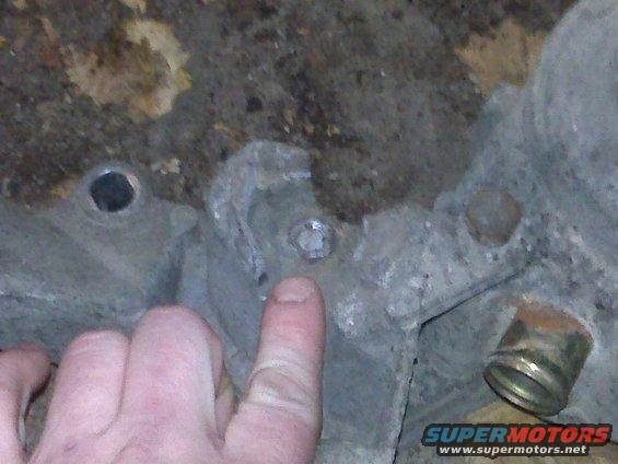 Close-up of a metal engine block with a bolt and a brass fitting. A finger points to the bolt