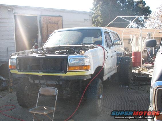 White Ford Bronco with hood open, missing grille, and yellow bumper trim. It's parked outside a garage