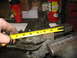 1995 Ford C-350 (Centurion Conversions) - A hand holds a tape measure against a worn suspension part secured in a vise, with tools and containers in the background.