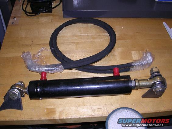 Hydraulic cylinder with two hoses, one coiled, on a wooden surface. Some parts are wrapped in plastic