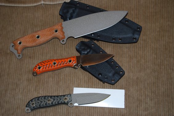 Three knives laid out on a brown corduroy surface, each with a black sheath