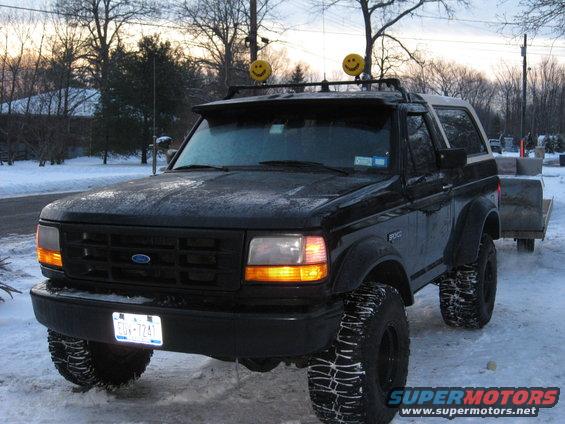 Black 1995 Ford Bronco with smiley face lights on roof rack, parked in snow, towing a trailer