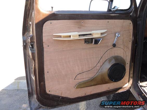 Brown car door interior with unfinished plywood panel, speaker cutout, and chrome door handle