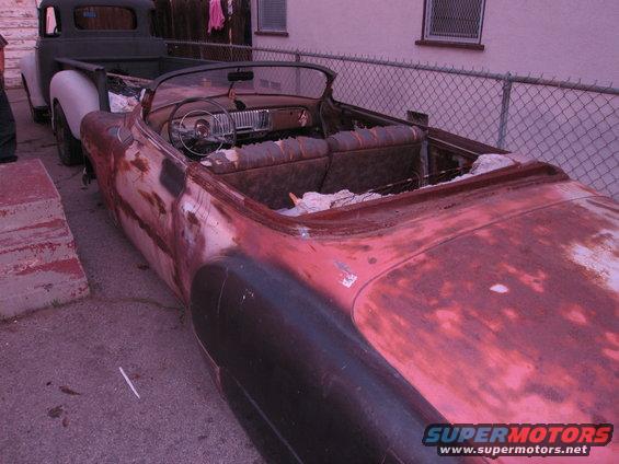 Rusty red convertible car interior with worn seats and dashboard, parked next to a pickup truck
