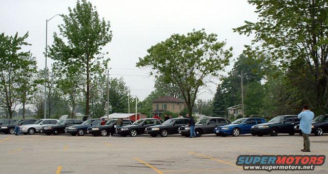Fleet of dark sedans, one blue sedan, parked in a lot. A man in a blue shirt stands near the right