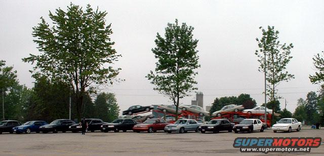 Several sedans are parked in a lot, with more cars stacked on car carriers in the background