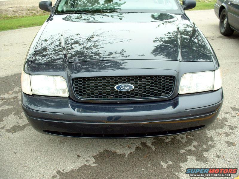 Front of a dark blue Ford Crown Victoria with a honeycomb grille and a Ford logo. Another vehicle is partially visible on the right