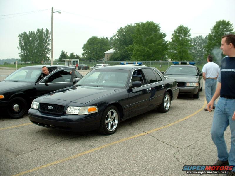Three black Ford Crown Victoria police cars parked in a lot, one with a man leaning out the driver's window