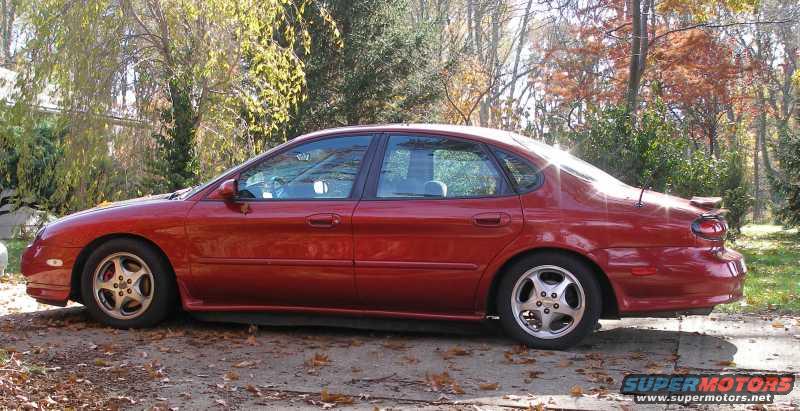 Red 1997 Ford Taurus sedan parked outdoors, side view, with autumn foliage in the background