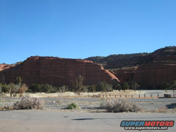 Desert landscape with red rock formations under a clear blue sky. Dry brush and scattered trees are in the foreground