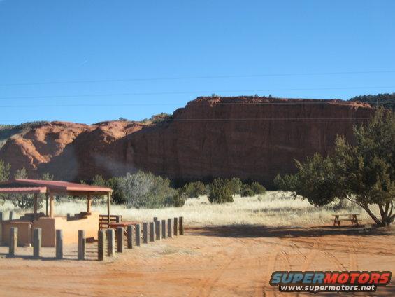 Desert landscape with red rock formations, a picnic shelter, and a picnic table under a tree. A "SUPERMOTORS.NET" watermark is visible