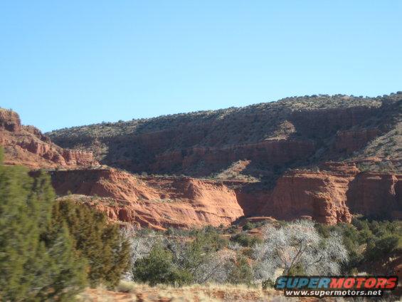 Desert landscape with red rock formations and sparse vegetation, Supermotors.net logo visible