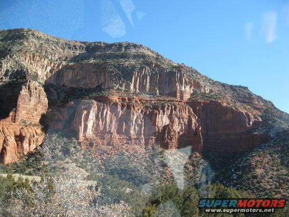 Red rock cliffs with sparse vegetation under a clear blue sky. A watermark reads SUPERMOTORS