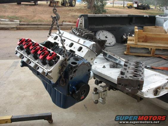 Engine block with red valve covers suspended by a chain, being loaded into a pickup truck bed with cylinder heads and other parts