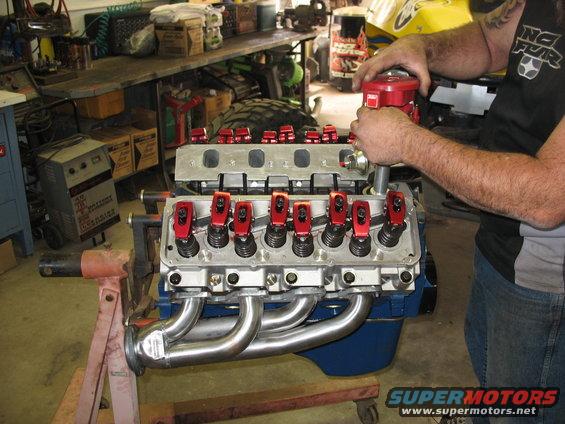 V8 engine with red valve covers and chrome headers on an engine stand, person installing distributor