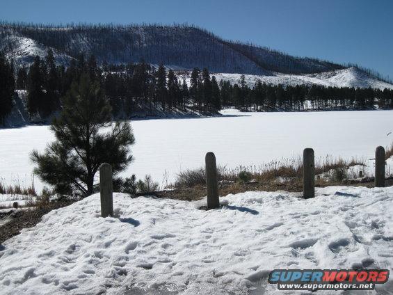 Snowy landscape with a frozen lake, pine trees, and a distant treed hill under a clear blue sky