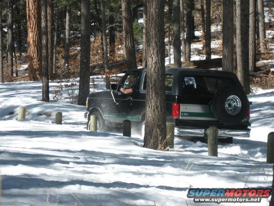 Black Ford Bronco with green stripe driving through snow, spare tire mounted on rear door