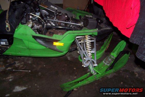 Green snowmobile with exposed engine and front suspension, resting on a wet floor