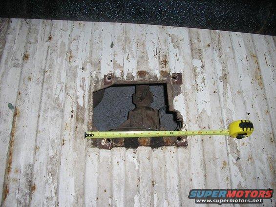 White corrugated metal surface with a square hole revealing rusty mechanical parts; yellow measuring tape across the hole