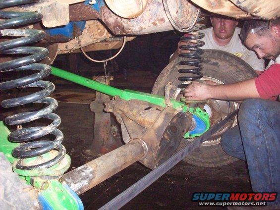 Mechanics work on the suspension of a vehicle, possibly a Ford Bronco, with large coil springs and green suspension components