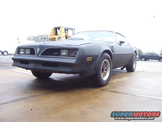 Black 1977 Pontiac Firebird with a scoop on the hood sits on concrete, yellow construction vehicle behind it