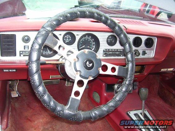 Interior shot of a 1978 Pontiac Trans Am with a black steering wheel and red dashboard. The bird emblem is on the steering wheel